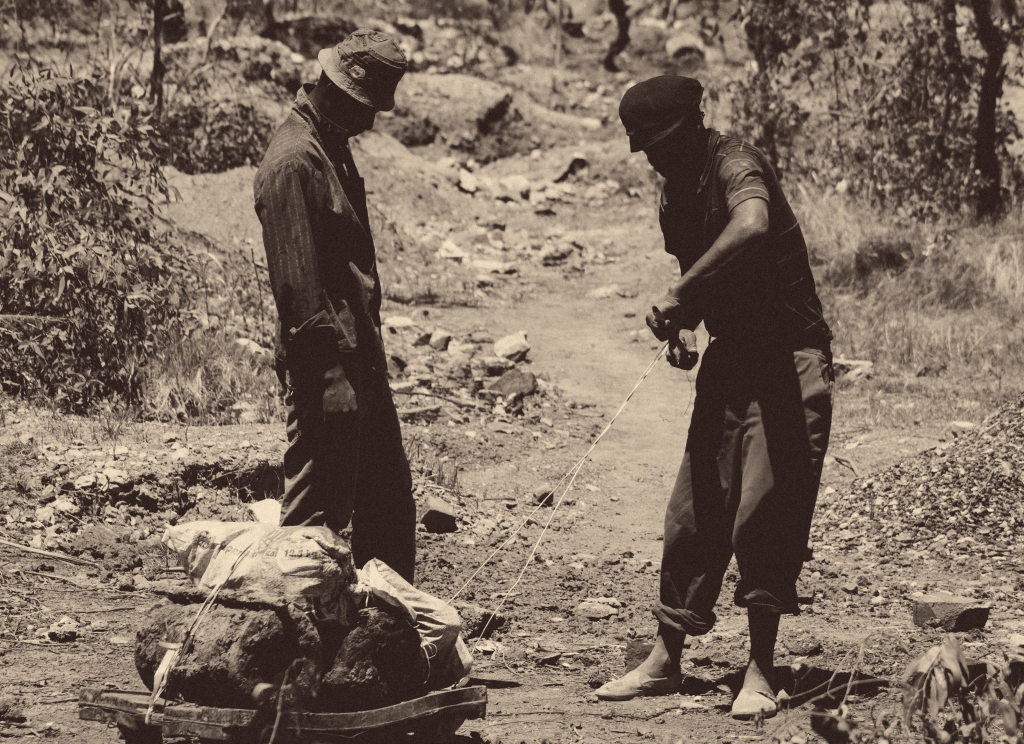Sepia-toned photo of 2 Black men dragging the scrap metal they collected on a makeshift trolley, untwisting the lines used to drag the trolley.